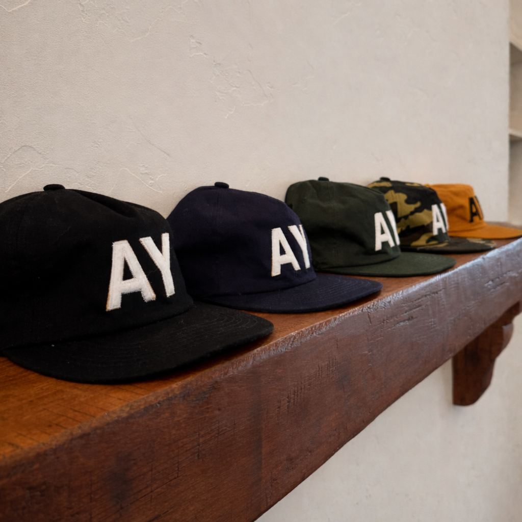 Collection of caps with 'AY' logo on a wooden shelf against a light wall.